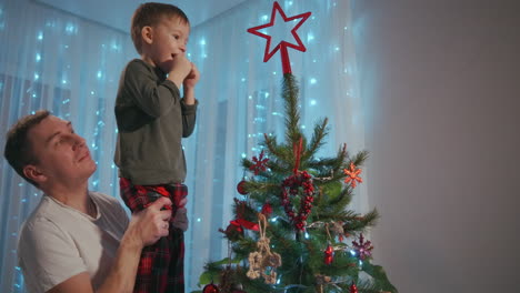 Dad-lifted-the-boy-to-the-top-of-the-Christmas-tree-to-place-a-Christmas-star-on-top-of-the-top.-Father-and-son-decorate-Christmas-tree-together.-High-quality-4k-footage